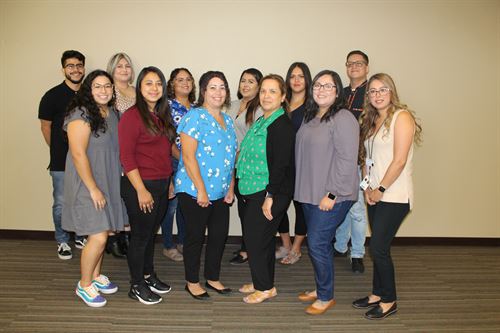 group of 12 women and men standing together for a group photo inside an office building with a just a blank wall as a backdrop.